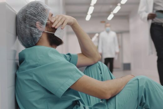 Exhausted and stressed medical worker in scrubs and safety mask resting on floor in hallway. Concept of is it harder to be a doctor or a lawyer